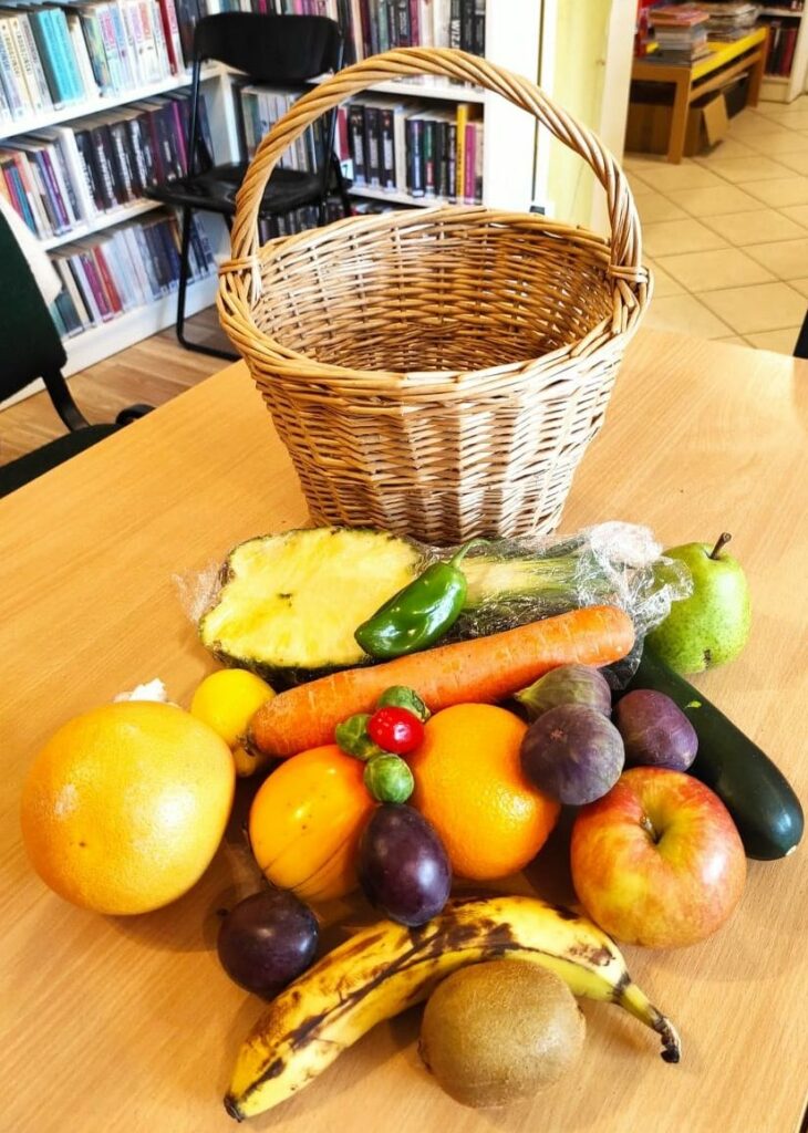 na zdjęciu widać stół na którym stoi wiklinowy kosz a przed nim poukładane są owoce i warzywa takie jak np. ananas, czosnek, cukinia, figi, banan, jabłko itp.