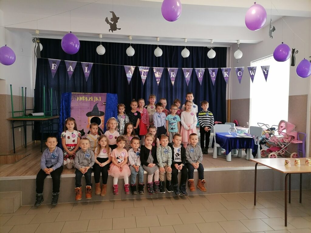 Na zdjęciu widoczna jest grupa 25 przedszkolaków pozujących do wspólnego zdjęcia. W tle foto-budka z czarownicą i zabawki. Nad głowami dzieci wisi napis "bal andrzejkowy".