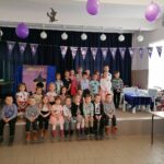 Na zdjęciu widoczna jest grupa 25 przedszkolaków pozujących do wspólnego zdjęcia. W tle foto-budka z czarownicą i zabawki. Nad głowami dzieci wisi napis "bal andrzejkowy".