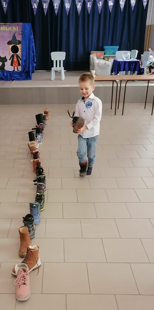 Na zdjęciu widoczny jest chłopiec trzymający w rękach swój but oraz powstały sznur z butów pozostałych przedszkolaków. W tle foto-budka z czarownicą oraz zabawki.