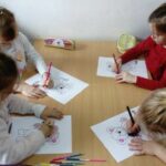 Przy małym stoliczku siedzą cztery dziewczynki i rysują kredkami na kartkach swoje ulubione bajkowe misie.