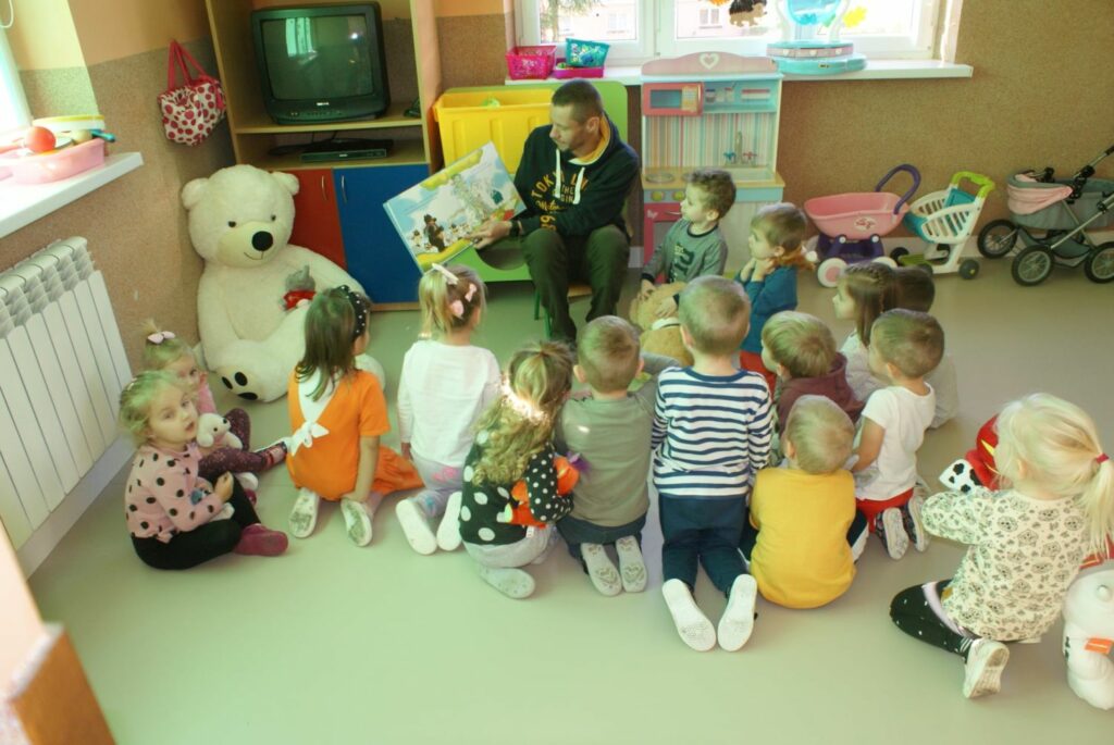 W jednej z przedszkolnych sal pan bibliotekarz siedzi na krześle i czyta bajkę o „Misiu – Tulisiu”, a wokół niego siedzą na podłodze zaciekawione dzieci.