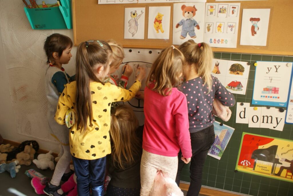 Na ścianie przypięto plakat wielkiego misia. Dookoła niego kłębią się przedszkolaki, które kolorują go kredkami.