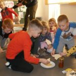 Na podłodze widzimy słoiczek i talerzyk z miodem wokół którego siedzą przedszkolaki i maczają w nim kawałki wafla, a następnie ze smakiem zjadają.
