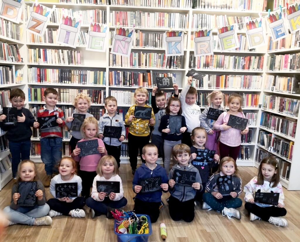 Na zdjęciu widać salę biblioteczną dzieci stoją przed regałami z książkami trzymają w rękach wykonane przez siebie obrazki. Nad nimi widać napis dzień kredki.