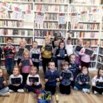 Na zdjęciu widać salę biblioteczną dzieci stoją przed regałami z książkami trzymają w rękach wykonane przez siebie obrazki. Nad nimi widać napis dzień kredki.