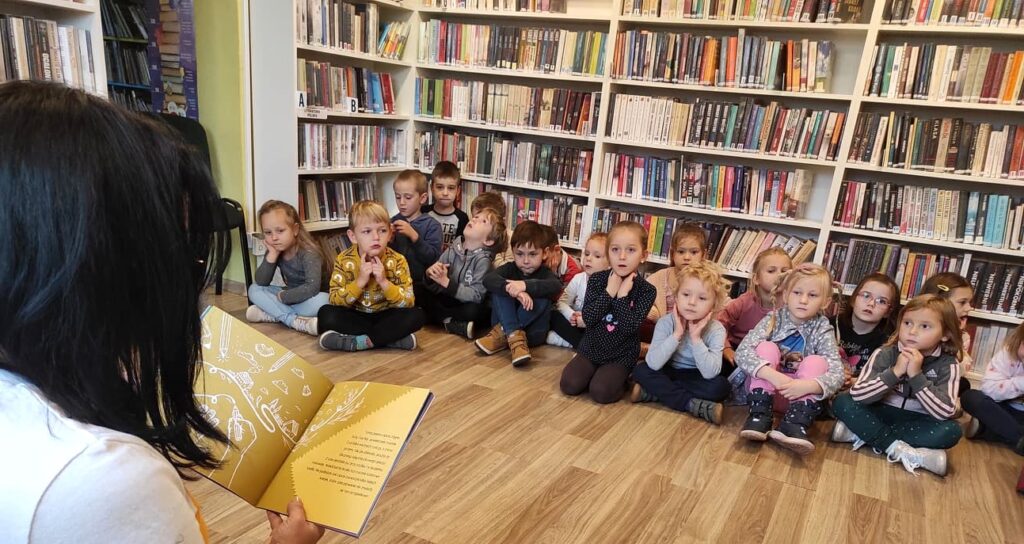 Na zdjęciu widać dzieci siedzące na podłodze i słuchające książki, która czytana im bibliotekarka.