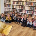 Na zdjęciu widać dzieci siedzące na podłodze i słuchające książki, która czytana im bibliotekarka.