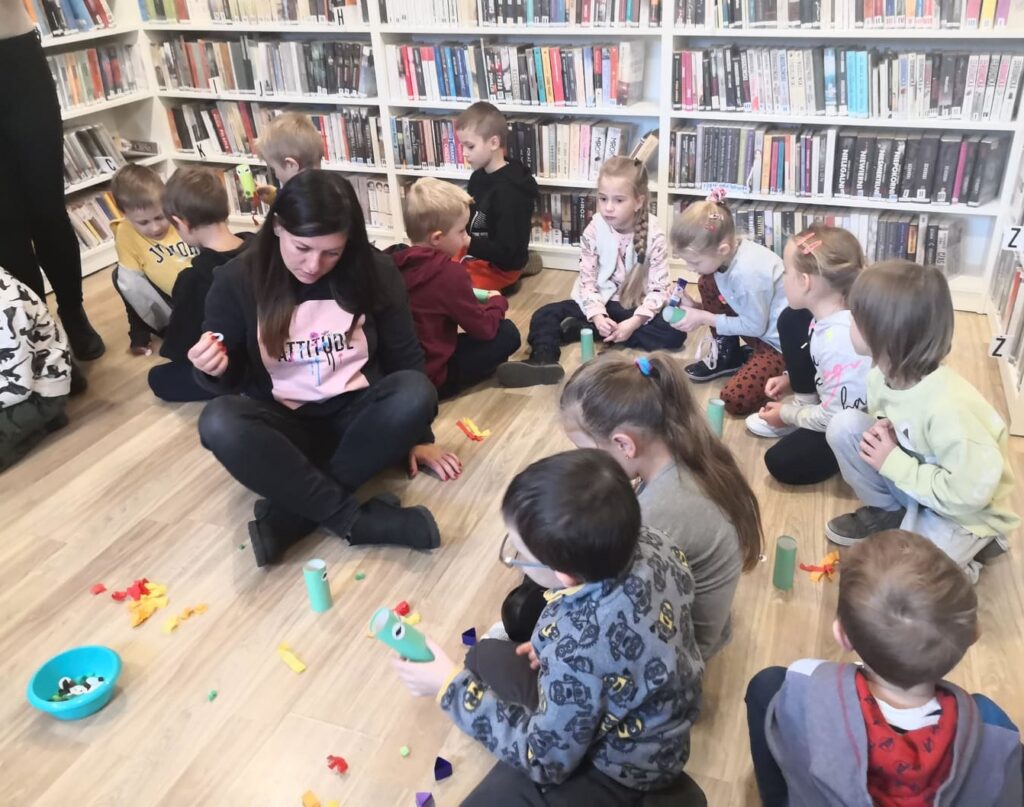 na zdjęciu widać bibliotekarkę siedząca po turecku na podłodze i pomagająca dzieciom w wykonaniu pracy plastycznej