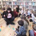 na zdjęciu widać bibliotekarkę siedząca po turecku na podłodze i pomagająca dzieciom w wykonaniu pracy plastycznej