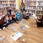 na zdjęciu widać bibliotekarkę oraz dzieci siedzące na podłodze. Przedszkolaki słuchając czytana bajkę. Bibliotekarka trzyma książkę . Na zdjęciu widać również regały z książkami.