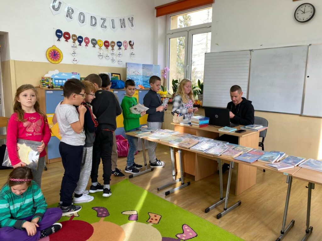 W szkolnej sali przy biurku siedzi bibliotekarz, przed nim na stolikach ułożonych jest mnóstwo kolorowych książek. Obok w długiej kolejce ustawili się uczniowie chcąc wypożyczyć nowe lektury.