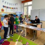 W szkolnej sali przy biurku siedzi bibliotekarz, przed nim na stolikach ułożonych jest mnóstwo kolorowych książek. Obok w długiej kolejce ustawili się uczniowie chcąc wypożyczyć nowe lektury.