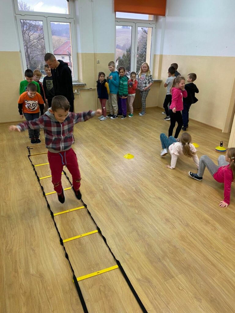 Mali sportowcy są ustawieni w kolejce w jednym końcu sali, przed nimi znajduje się ułożony z drabinki koordynacyjnej i stożków sprawnościowy tor przeszkód, który każdy musi pokonać.