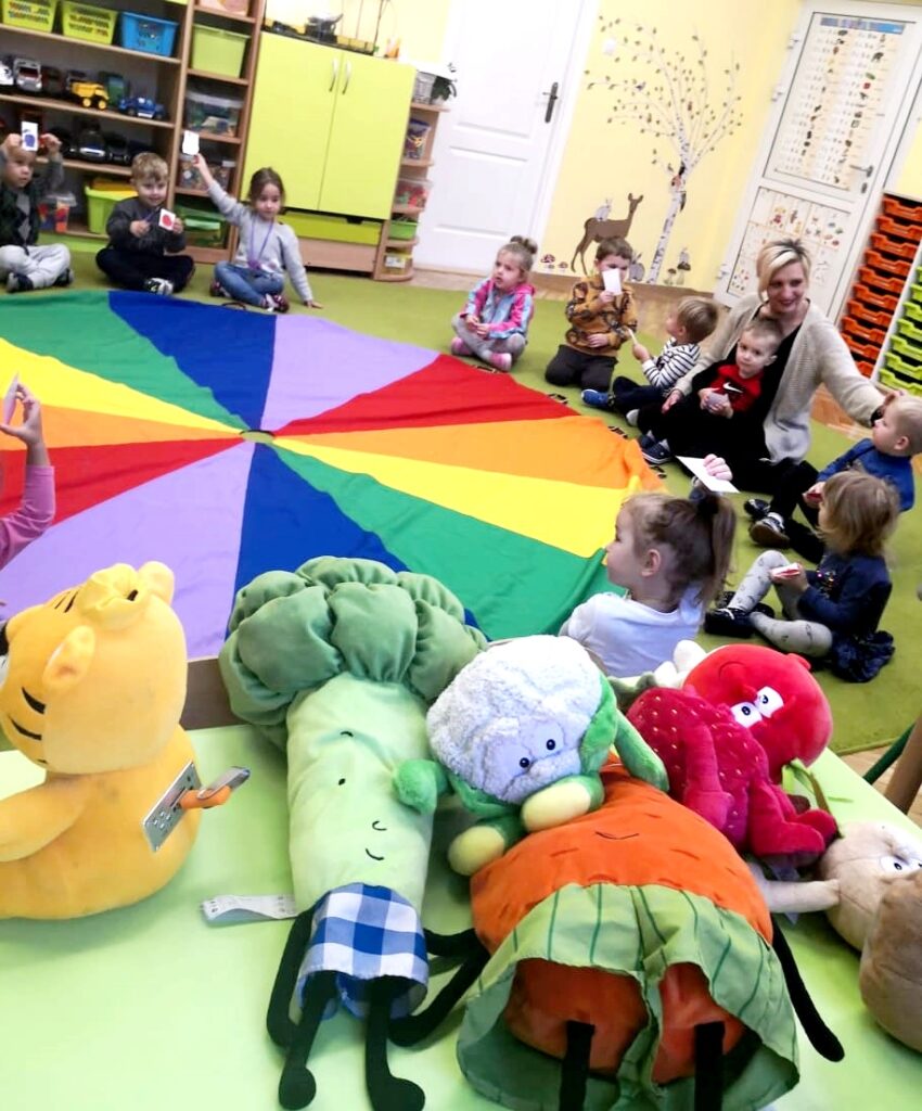 Na ziemi wokół chusty klanzy siedzą dzieci z ochronki wraz z panią wychowawczynią. Na pierwszyn planie pluszowe owoce i warzywa