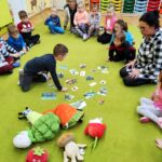 Na jasno zielonej wykładzinie siedzi pani bibliotekarka oraz dzieci z ochronki . Na kolorowych obrazkach jeden z chłopców rozpoznaje warzywa i owoce . Obok na ziemi leżą pluszowe owoce i warzywa