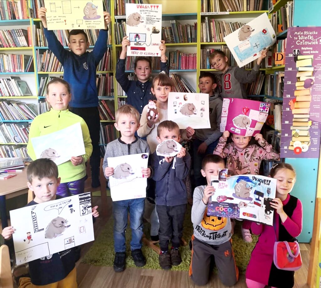 Na fotografii widać sale biblioteczną. Na tle regałów stoją dzieci trzymając kolorowe plakaty wykonane przez siebie.