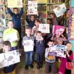 Na fotografii widać sale biblioteczną. Na tle regałów stoją dzieci trzymając kolorowe plakaty wykonane przez siebie.