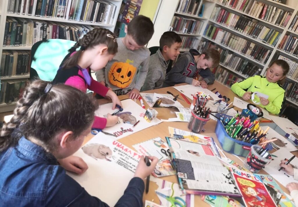 Na fotografii widać dzieci które siedzą wokół stołu wykonując kolorowe prace plastyczne. Stół zapełniony jest różnego rodzaju materiałami plastycznymi.