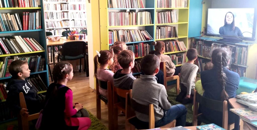Na zdjęciu widać dzieci siedzące i oglądające telewizję w sali bibliotecznej w której widoczne są również regały z książkami.