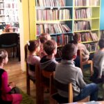 Na zdjęciu widać dzieci siedzące i oglądające telewizję w sali bibliotecznej w której widoczne są również regały z książkami.