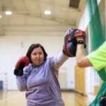 Na zdjęciu widoczna jest jedna z uczestniczek projektu "MAMY-to!" oraz trener podczas zajęć sportowych na sali gimnastycznej. Oboje ubrani są w stroje sportowe. Kobieta na rękach ma założone rękawice bokserskie i uderza w worek treningowy. Trener instruuje uczestniczkę zajęć jak prawidłowo wykonywać uderzenia. W tle worek treningowy oraz drabinki.