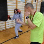 Na zdjęciu widoczna jest jedna z uczestniczek projektu "MAMY-to!" oraz trener podczas zajęć sportowych na sali gimnastycznej. Oboje ubrani są w stroje sportowe. Kobieta na rękach ma założone rękawice bokserskie a trener tarcze do sparingu. W tle worek treningowy oraz drabinki.