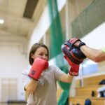 Na zdjęciu widoczna jest jedna z uczestniczek projektu "MAMY-to!" oraz trener podczas zajęć sportowych na sali gimnastycznej. Oboje ubrani są w stroje sportowe. Kobieta na rękach ma założone rękawice bokserskie a trener tarcze do sparingu.