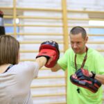 Na zdjęciu widoczna jest jedna z uczestniczek projektu "MAMY-to!" oraz trener podczas zajęć sportowych na sali gimnastycznej. Oboje ubrani są w stroje sportowe. Kobieta na rękach ma założone rękawice bokserskie a trener tarcze do sparingu.