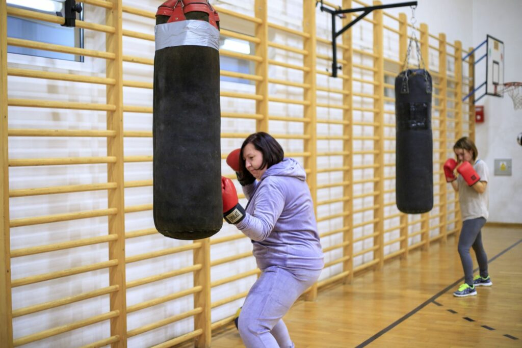 Na zdjęciu widoczne są 2 dwie uczestniczki projektu "MAMY-to!" podczas zajęć sportowych na sali gimnastycznej. Uczestniczki ubrane są w stroje sportowe, na rękach mają założone rękawice bokserskie i uderzają w worki treningowe.