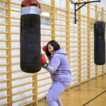 Na zdjęciu widoczne są 2 dwie uczestniczki projektu "MAMY-to!" podczas zajęć sportowych na sali gimnastycznej. Uczestniczki ubrane są w stroje sportowe, na rękach mają założone rękawice bokserskie i uderzają w worki treningowe.
