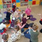 na zdjęciu widać dzieci siedzące na podłodze za nimi widać kolorowe szafki. Przed dziećmi siedzi bibliotekarka w czerwonym swetrze.