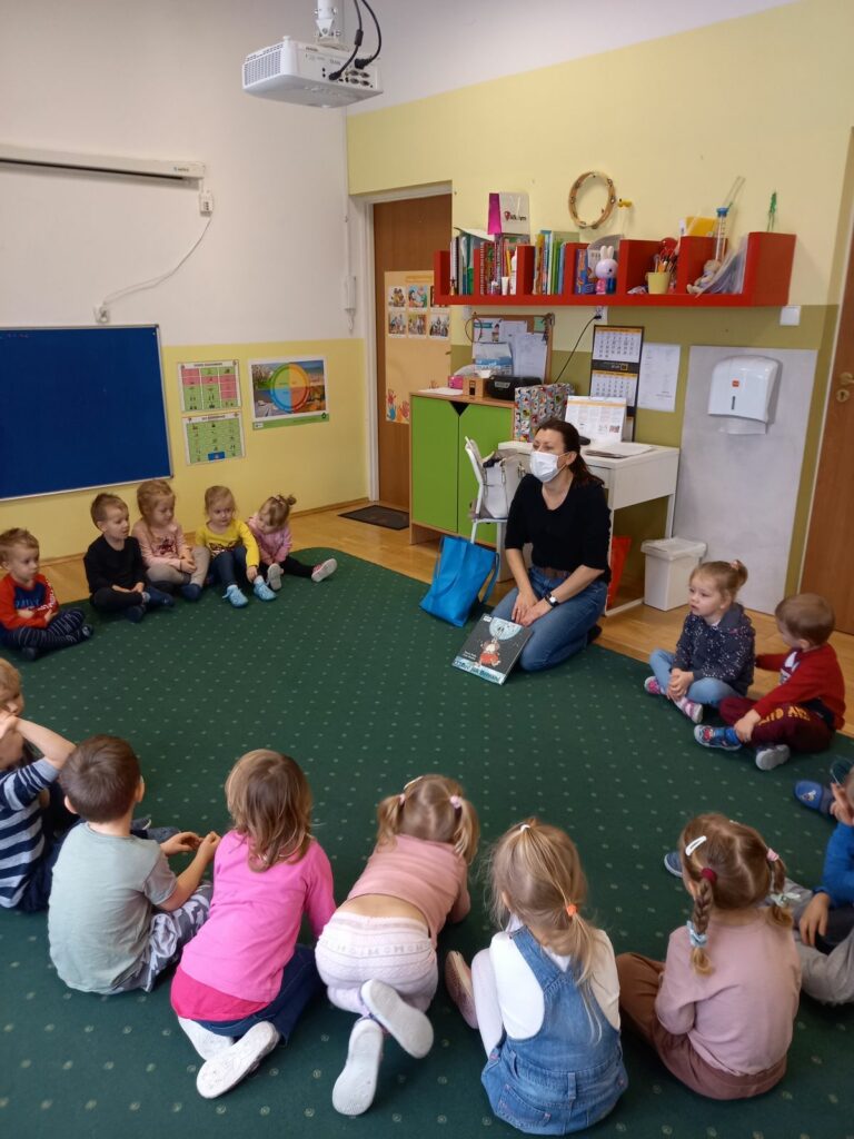 Na ziemi w okręku siedzą dzieci wśród nich jest pani bibliotekarka, która prezentuje przyniesioną książkę.