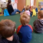 Na ziemi siedzi pani bibliotekarka oraz przedszkolaki. Pani bibliotekarka trzyma w ręku książkę i pokazuje ilustracje.