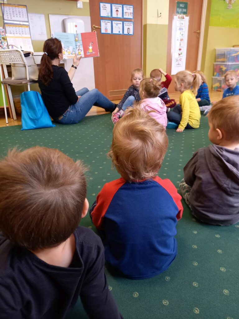 Na ziemi siedzi pani bibliotekarka oraz przedszkolaki. Pani bibliotekarka trzyma w ręku książkę i pokazuje ilustracje.