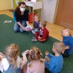 Na ziemi siedza dzieci oraz pani bibliotekarka, która częstuje dzieci cukierkami.