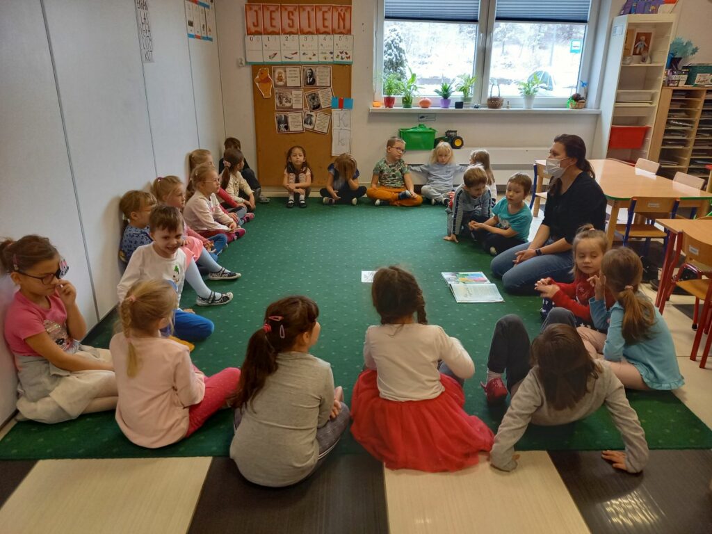 Na ziemi w okręgu siedzą przedszkolaki oraz pani bibliotekarka, która zadaje pytania dzieciom.