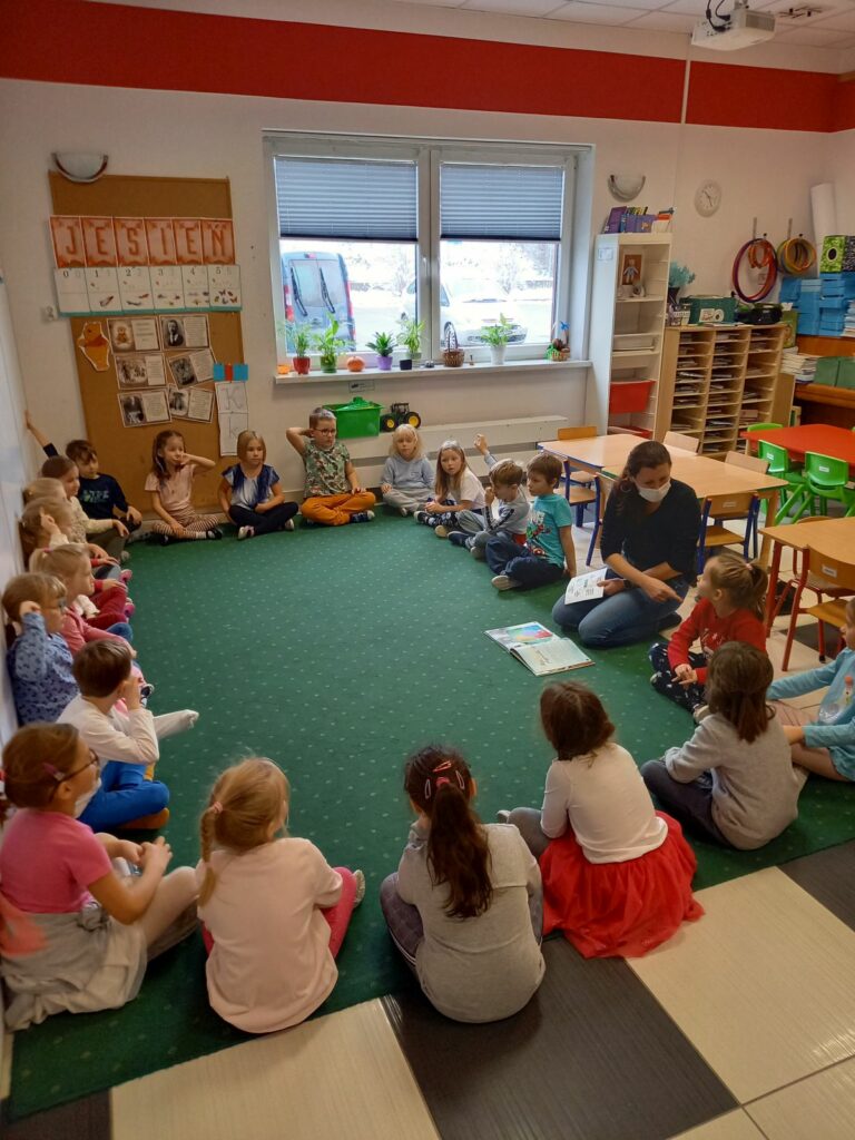 Na ziemi w okręgu siedzą przedszkolaki oraz pani bibliotekarka, która zadaje pytania dzieciom.