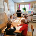 W bibliotecznej sali grupa dzieci siedzi przy długim stole i z zainteresowaniem słucha czytanej im przez bibliotekarza książki.