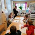 Siedzące przy stoliku dzieci z uwagą oglądają pokazywaną im przez bibliotekarza ilustrację z książki „Święta na świecie”.