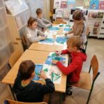 Mali artyści pracują w skupieniu przy stole wycinając, klejąc i kolorując swoje prace, z których powstaną niepowtarzalne choinki.