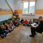 W klasie szkolnej pan bibliotekarz siedzi na krześle i czyta siedzącym wokół niego uczniom śląskie legendy.