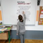 Jeden z chłopców stoi przy tablicy i zielonym flamastrem wpisuje do znajdującej się tam wielkiej krzyżówki odgadnięte przez siebie hasło.