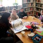 Przy stole siedzi pani bibliotekarka i czyta trzem chłopcom i jednej dziewczynce bajkę. Za dziećmi półki z książkami