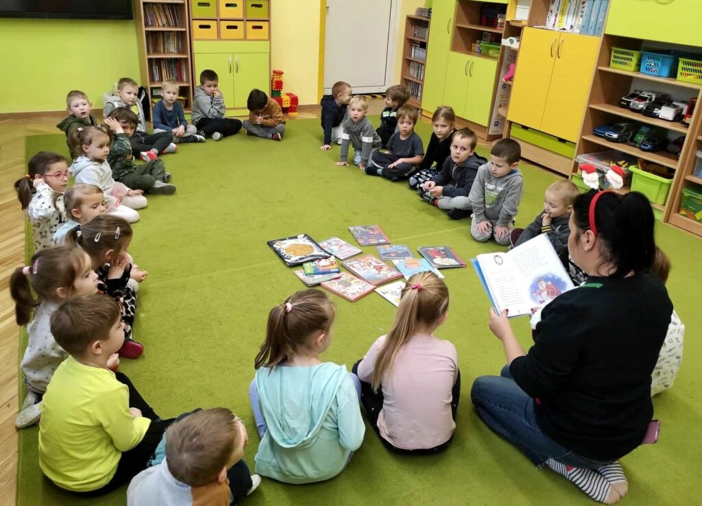Na zielonej wykładzinie w okręgu siedzą dzieci i pani bibliotekarka, która czyta dzieciom ksiażkę.
