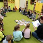 Na zielonej wykładzinie w okręgu siedzą dzieci i pani bibliotekarka, która czyta dzieciom ksiażkę.