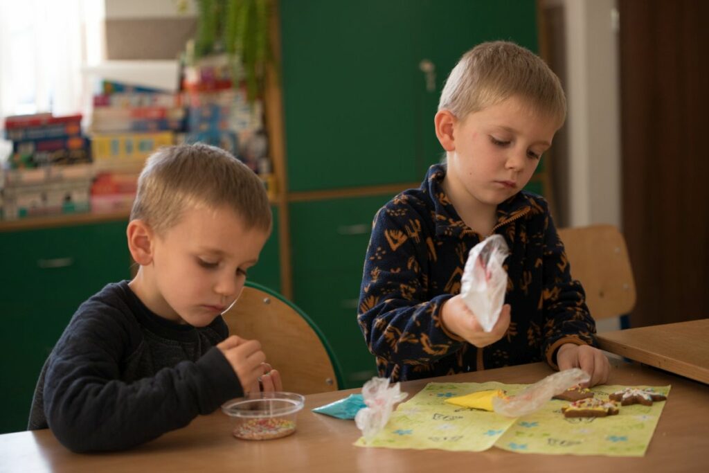 Na zdjęciu widocznych jest dwóch chłopców podczas dekorowania pierniczków. Obaj chłopcy ubrane mają ciemne bluzy. W tle zielone szafki z grami planszowymi.