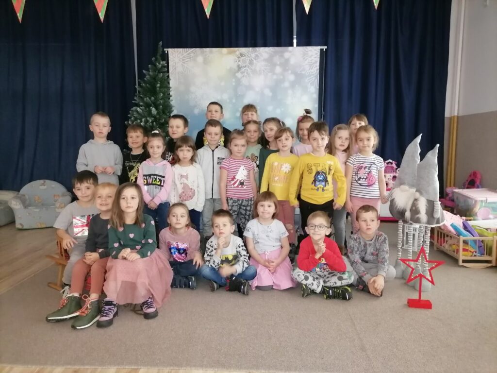 Na zdjęciu pozuje grupa dzieci przedszkolnych w wielokolorowych ubraniach. Z tyłu za nimi granatowe zasłony.