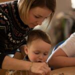 Na zdjęciu widoczna jest mama z dzieckiem dekorujący pierniczki. Kobieta ubrana jest w ciemny sweter z świątecznymi wzorami, chłopiec ubraną ma koszulę w kratę w odcieniach brązu i bieli.