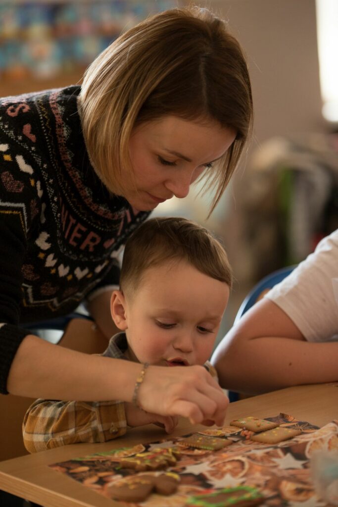 Na zdjęciu widoczna jest mama z dzieckiem dekorujący pierniczki. Kobieta ubrana jest w ciemny sweter z świątecznymi wzorami, chłopiec ubraną ma koszulę w kratę w odcieniach brązu i bieli.